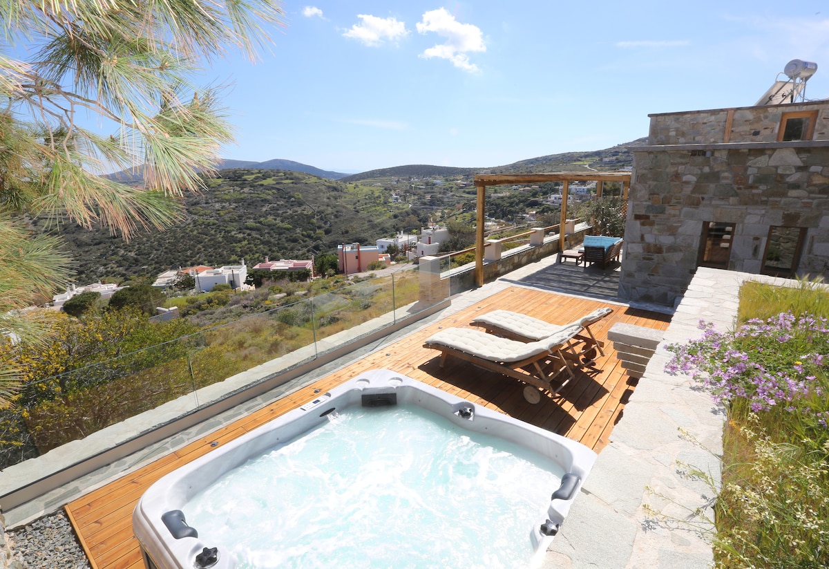 A spacious outdoor area features a large hot tub surrounded by wooden decking, with a sun lounger positioned nearby. Panoramic views of the surrounding hills and distant buildings are visible, complemented by the clear blue sky.
