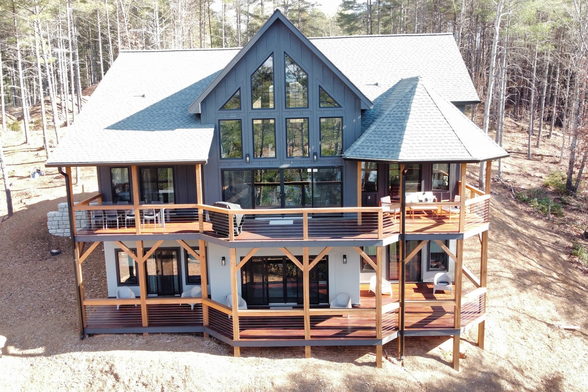The exterior of a modern rustic home is captured from above, showcasing large windows and multiple decks. The structure is surrounded by trees, with ample outdoor seating visible on the lower-level deck. The multi-gabled roof features a central peak that enhances the architectural design.