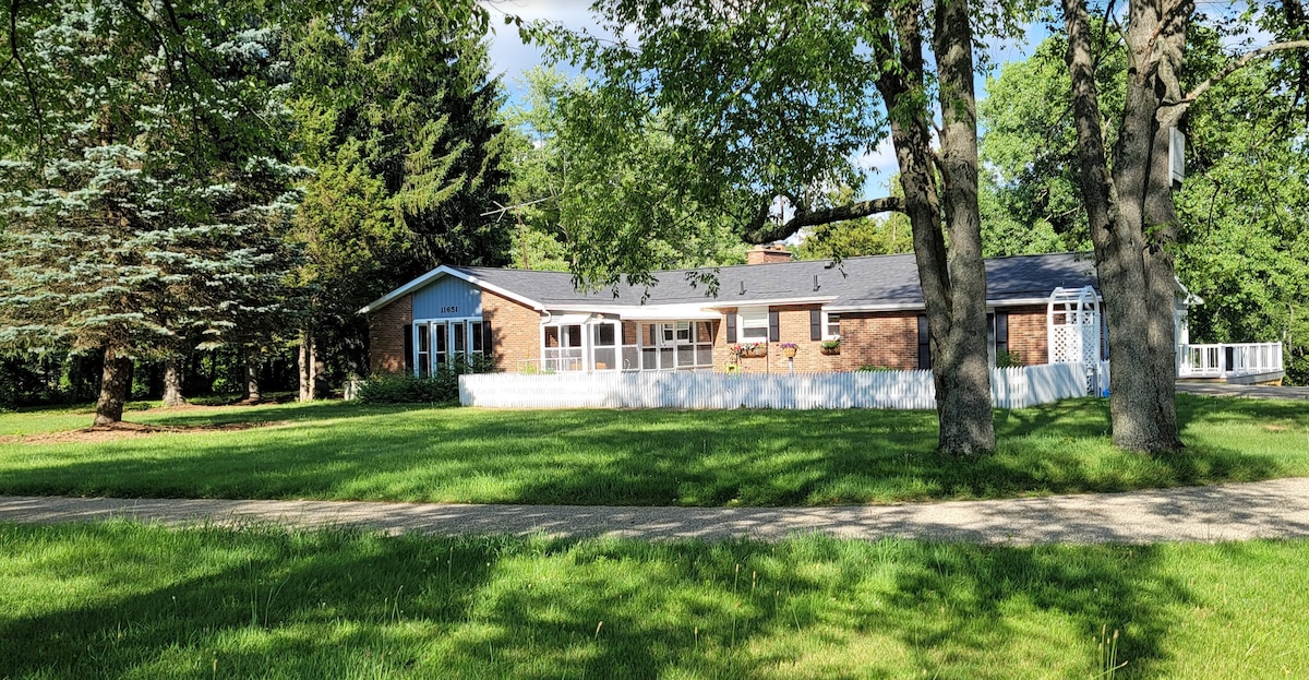 The spacious exterior view of the family estate showcases a brick structure surrounded by lush green lawns and mature trees. Natural light illuminates the building, highlighting large windows and a welcoming front porch. A gravel pathway leads gently towards the home.