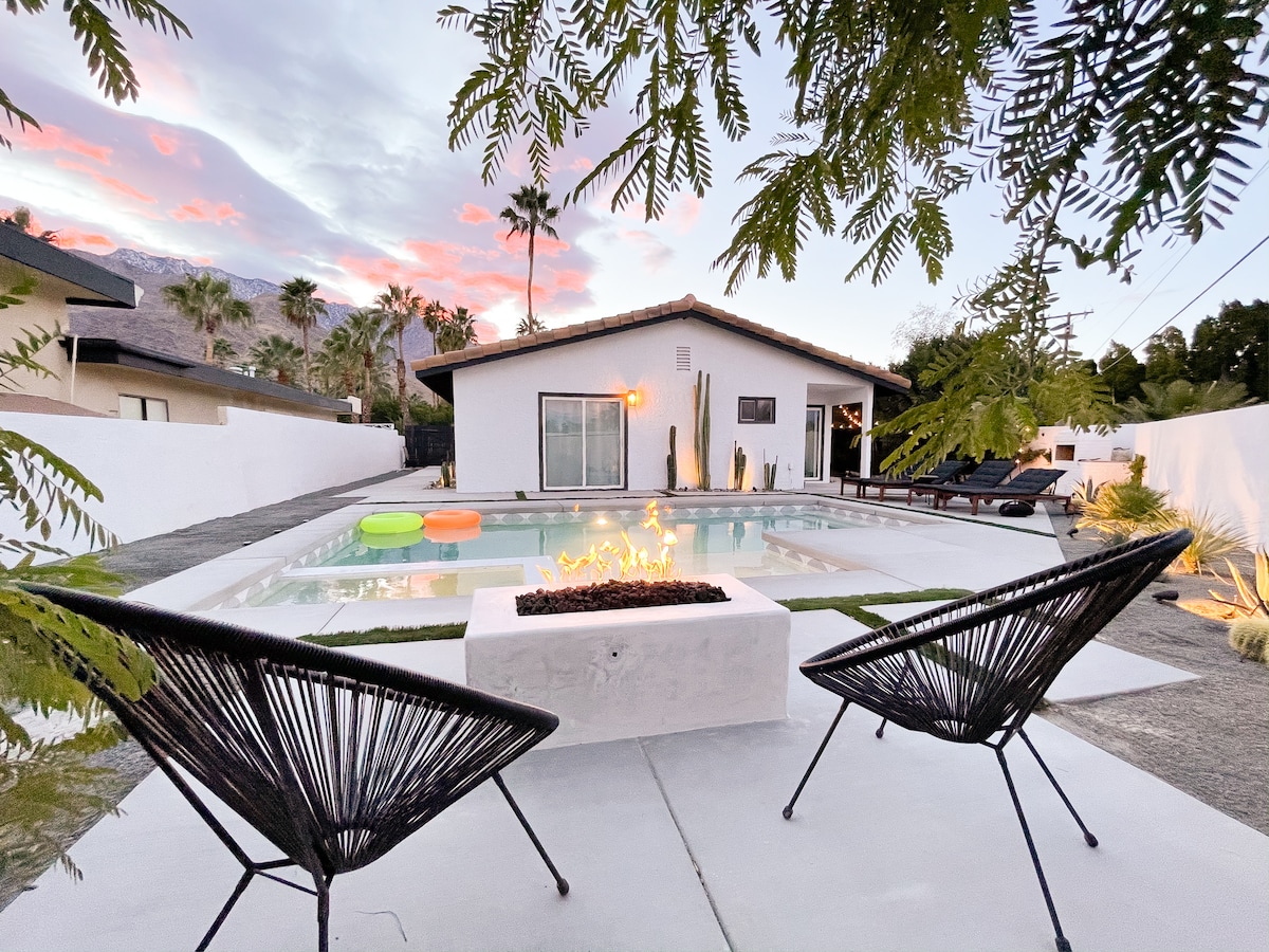 A serene backyard is highlighted by a sparkling saltwater pool surrounded by modern lounge chairs. A fire pit with flickering flames sits between two black chairs, while palm trees frame the scene against a colorful sunset sky.