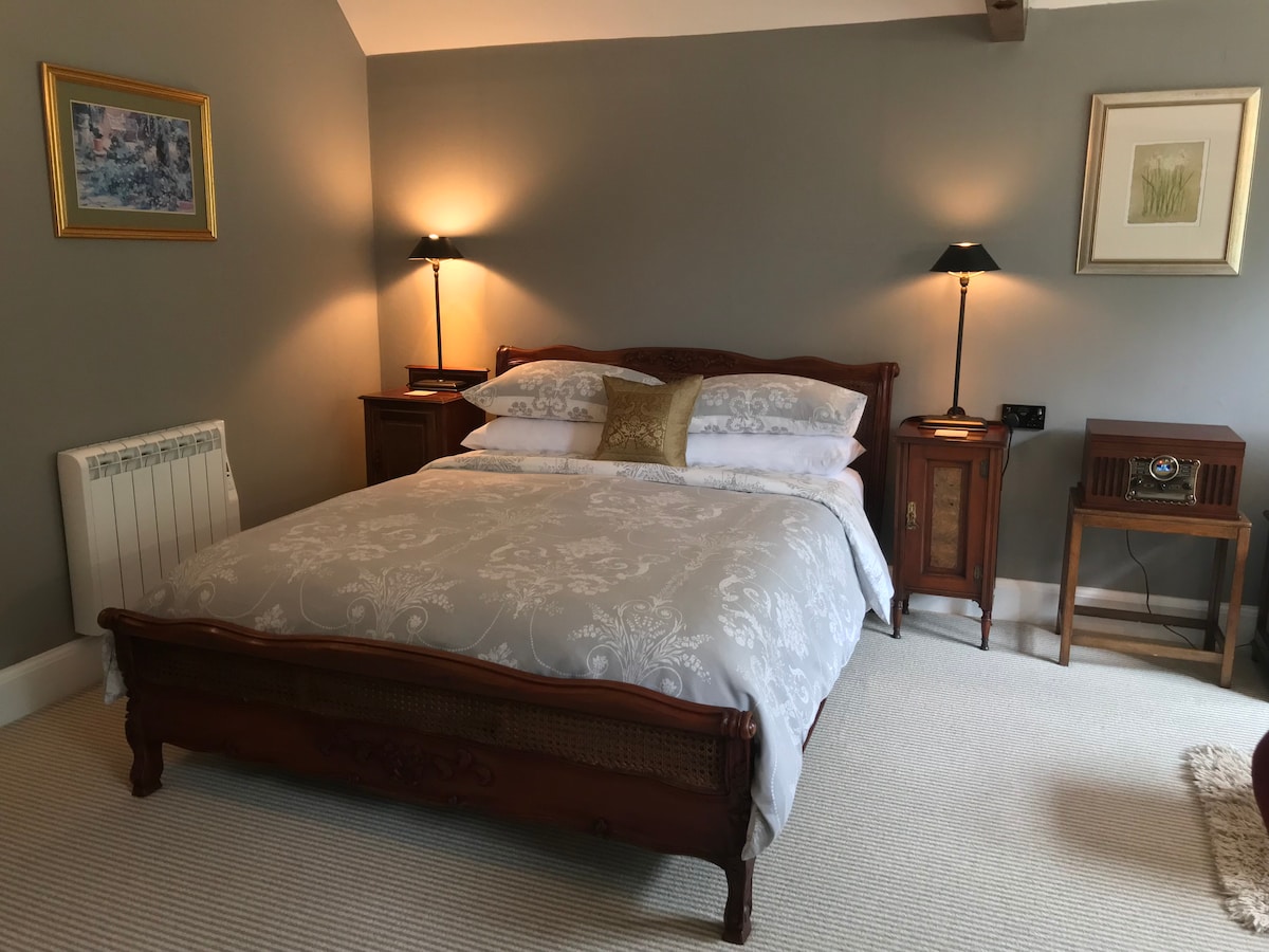 The bedroom features a wooden king-size bed dressed in light linens, complemented by two nightstands with lamps. A vintage radio sits against one wall, with soft, neutral-toned walls and carpet enhancing the serene environment.