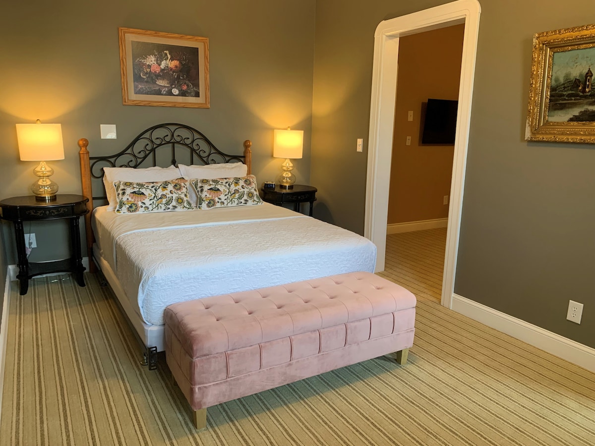 A queen-sized bed is placed against a neutral wall, flanked by two bedside tables with lamps. A decorative bench in soft pink sits at the foot of the bed. Artwork adorns the walls, contributing to the inviting ambiance of the room.