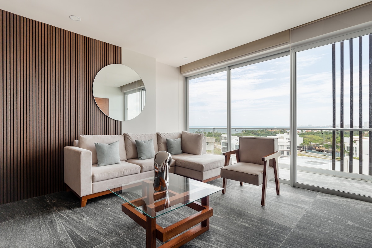 A bright living area features a modern sofa with soft cushions and a glass coffee table. A large mirror is mounted on a textured wooden wall. Floor-to-ceiling windows allow natural light to flood the space, while offering views of the surrounding area.