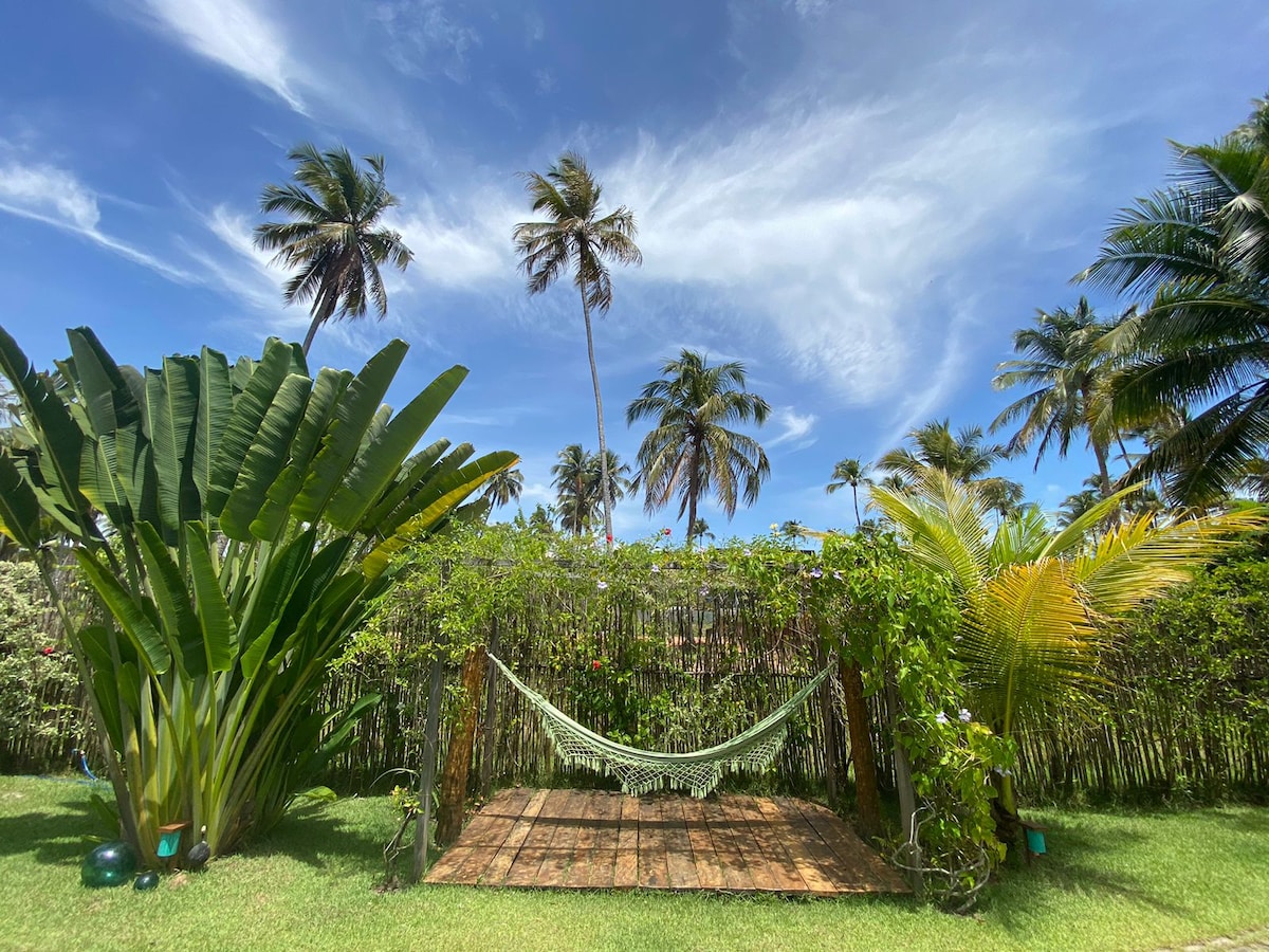 A serene outdoor space is framed by tall palm trees and lush greenery. A wooden deck features a comfortable hammock surrounded by vibrant tropical plants, inviting relaxation under a bright blue sky with wispy clouds.