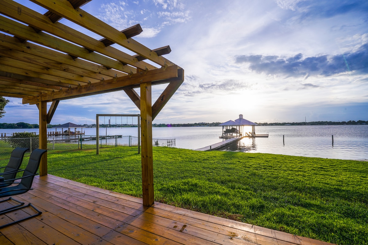 A spacious deck is seen beneath a wooden pergola, showcasing a view of Lake Clay. The grassy area extends toward the water, highlighting the private boat dock in the background. Soft light reflects off the lake's surface as the sun sets.