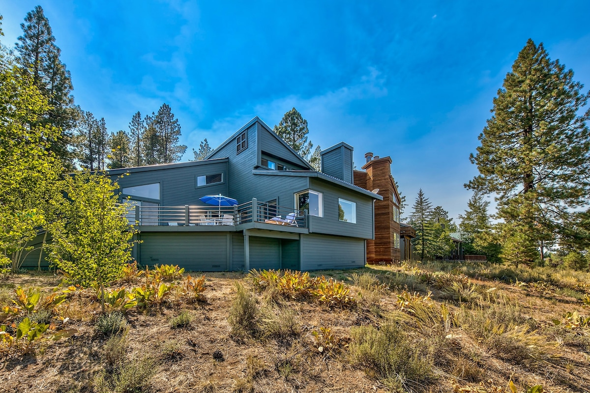 The exterior of the home showcases a contemporary design with large windows, surrounded by a natural landscape featuring tall trees and lush greenery. A deck extends from the house, furnished with outdoor seating and an umbrella for shade.