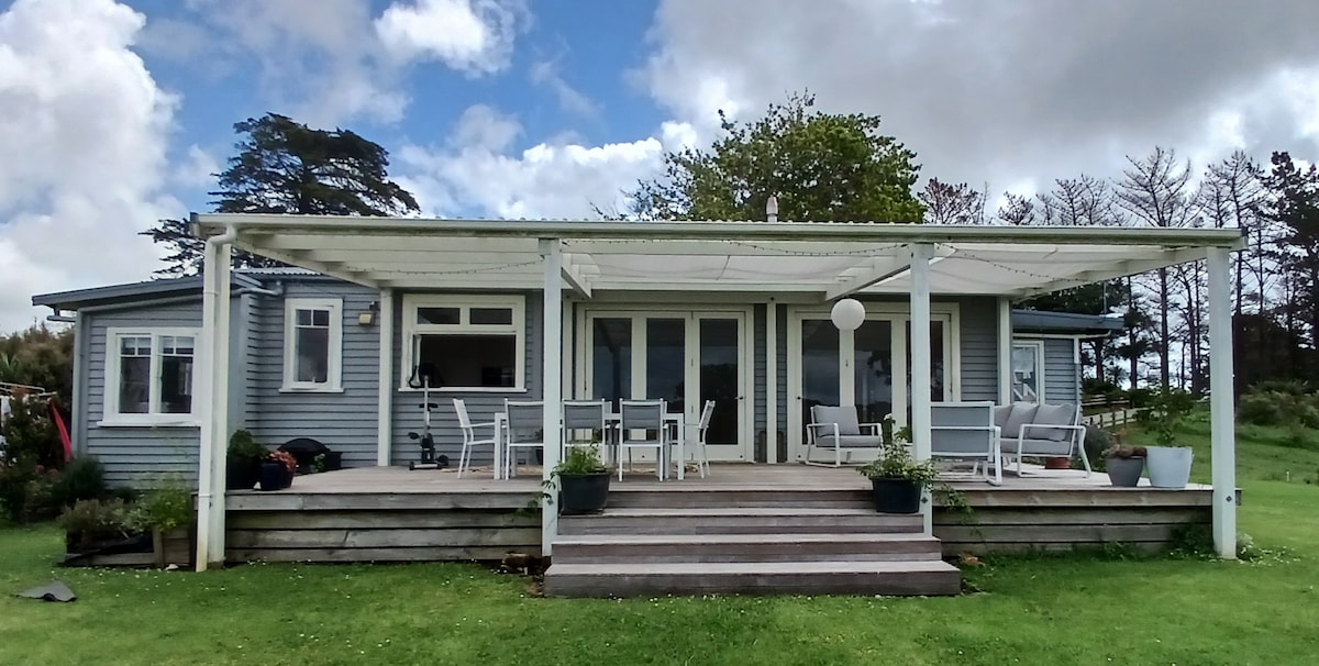 A renovated bungalow is shown with a large deck area, furnished with multiple outdoor chairs and tables. A covered seating space provides shade, alongside potted plants. The exterior features light blue siding, and large glass doors allow natural light to enter the interior.