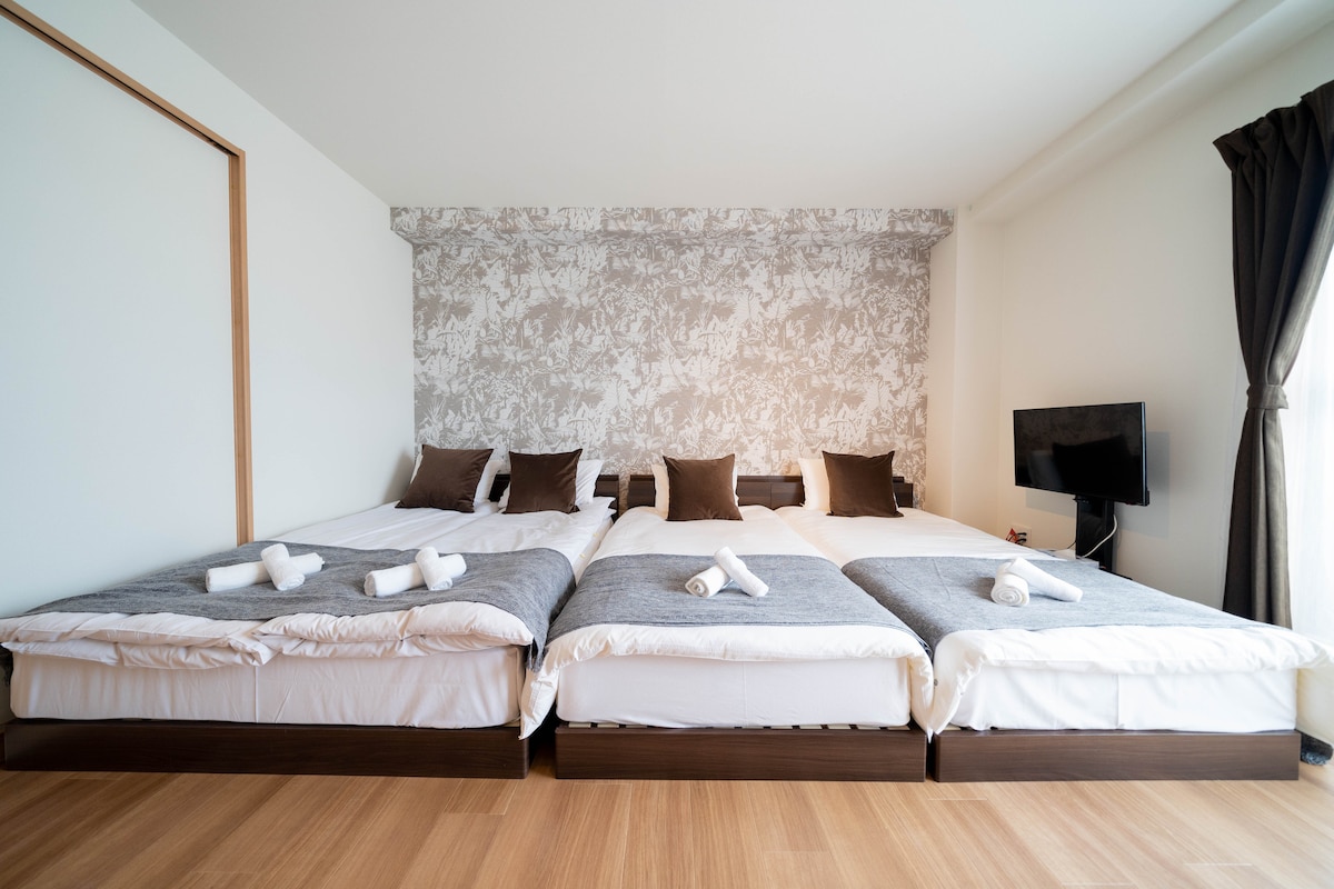 A spacious sleeping area is furnished with multiple beds arranged in a line, each featuring crisp white linens and light gray throws. Brown accent pillows add a touch of comfort. A television is mounted on the wall opposite, with natural light streaming in from a large window.