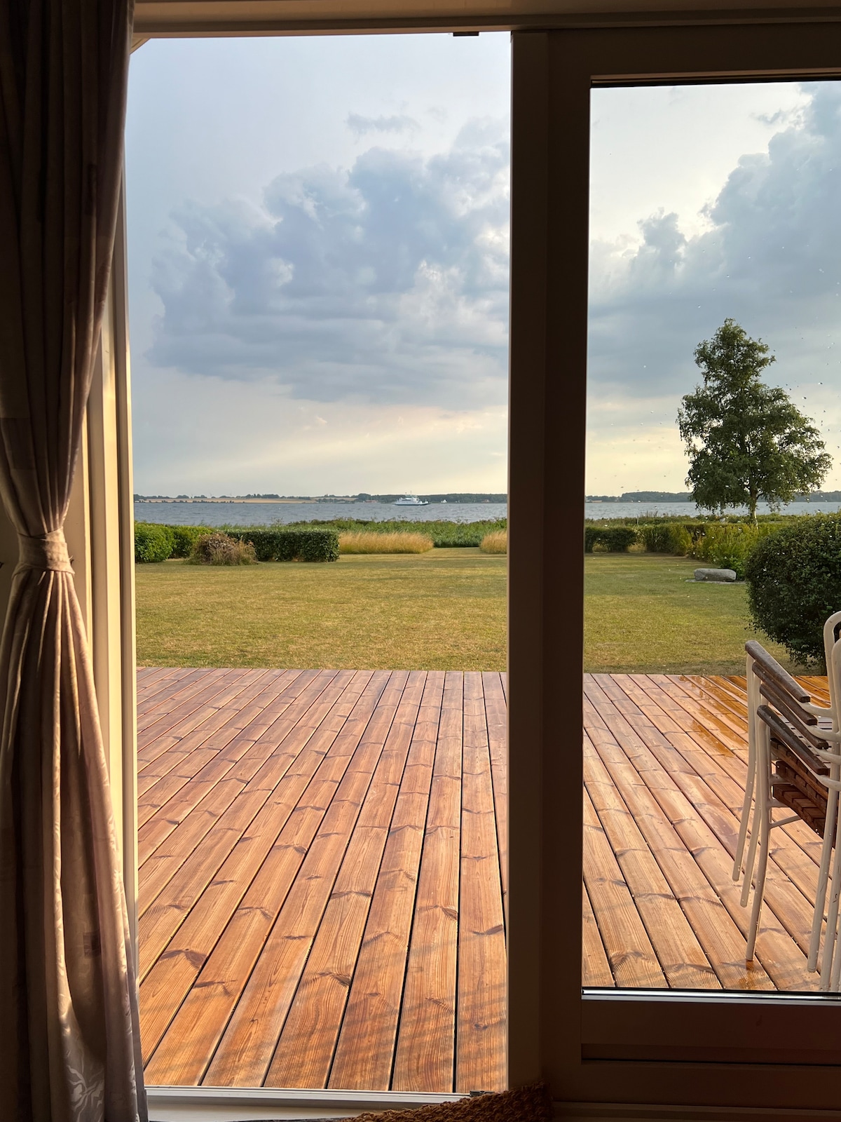 A view through large glass doors reveals a vibrant garden and wooden terrace. Lush green grass extends towards the horizon, framed by puffy clouds in a gray sky. A table and chairs are positioned on the terrace, inviting relaxation.