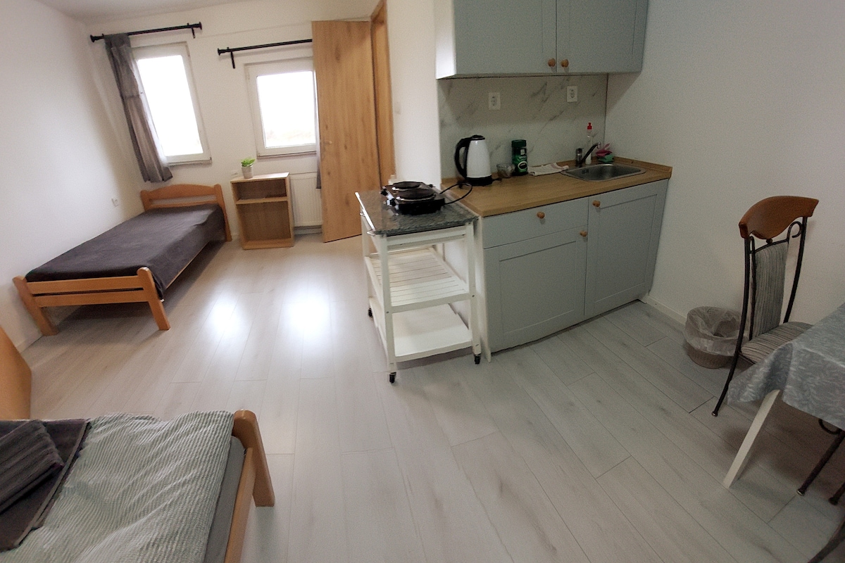 The studio features a functional kitchen area with a wooden countertop and essential appliances. Two single beds are present, with one partially covered by a gray blanket. A small dining table and a chair complement the space, while natural light enters through window curtains.