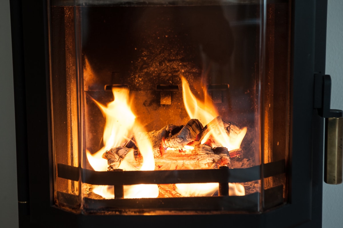 A wood-burning stove is depicted, with flames dancing over burning logs. The glass front offers a clear view of the fire, which casts a warm glow across the surrounding area. Charred wood and ashes are visible at the base.