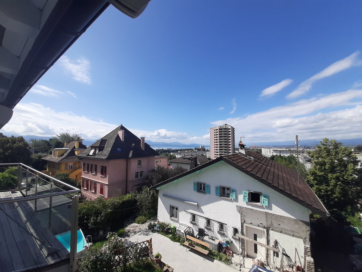 A view overlooking the neighborhood is presented, featuring a mix of colorful buildings and greenery. The horizon shows distant mountains under a clear blue sky, while a small flower garden and outdoor seating are visible in the foreground.