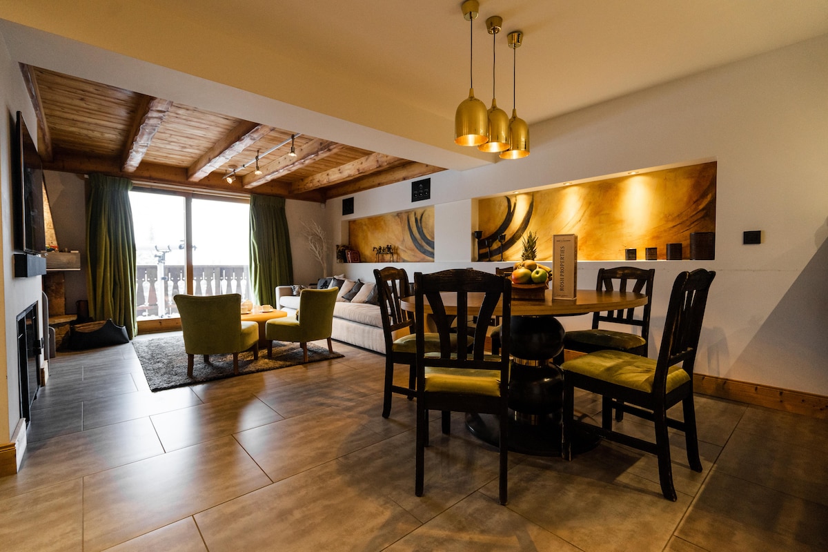 The living area features a spacious seating arrangement with two green chairs and a light-colored sofa, complemented by a round dining table surrounded by black chairs. Natural light flows in through large windows, with warm wooden beams accentuating the ceiling.
