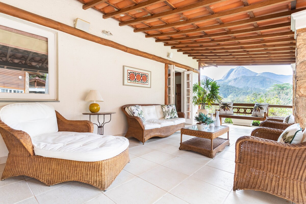 A covered outdoor seating area features comfortable wicker furniture, including a spacious sofa and armchairs, arranged around a wooden coffee table. Lush greenery and a scenic mountain view are visible in the background, creating a serene space for relaxation.