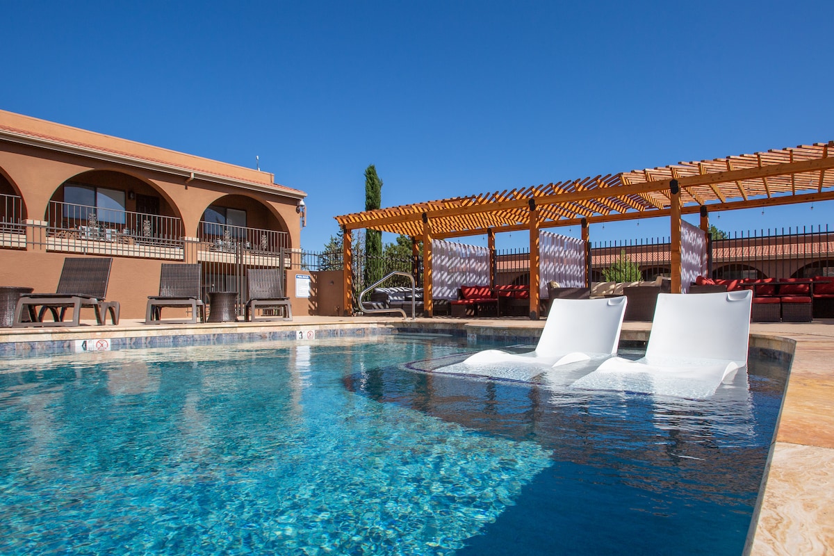 The outdoor pool features clear blue water with sun loungers partially submerged, creating a relaxing space. Surrounding the pool are shaded cabanas and lounge areas, set against a backdrop of the building's warm tones and blue sky.