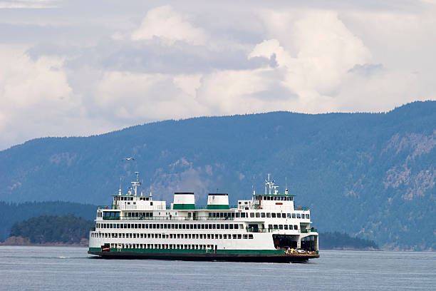 A large ferry is seen traveling across the water, surrounded by lush green hills. The ferry's distinct white and green design contrasts against the serene backdrop of the mountains and clouds, highlighting its connection to the nearby ferry terminal.