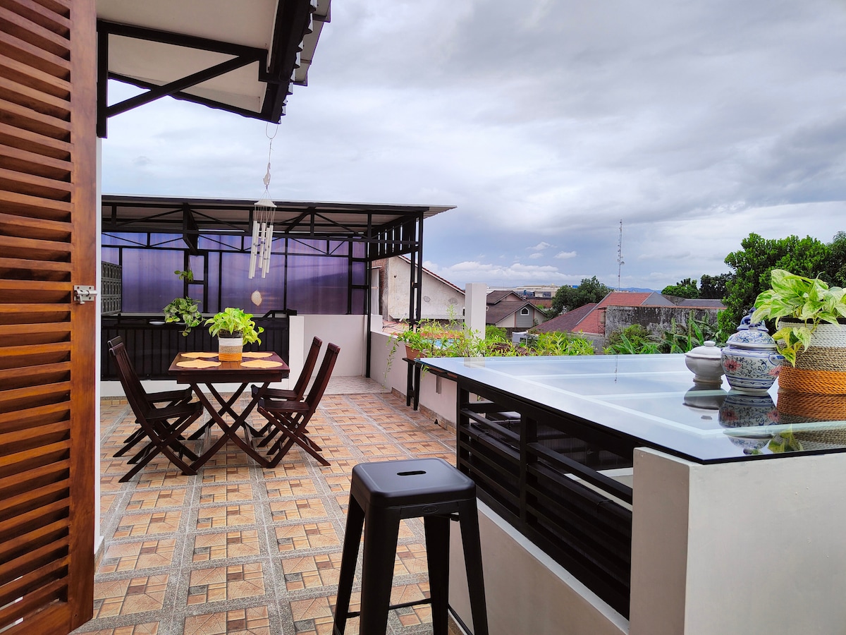An outdoor terrace space features a wooden dining set accompanied by potted plants. A sleek countertop with chairs offers a spot for relaxation, while large windows allow natural light to fill the area. Scenic views of the surrounding neighborhood are visible.