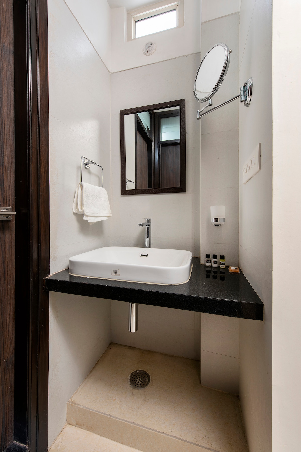 A modern en-suite bathroom is designed with sleek, minimalistic fixtures. A compact sink is mounted atop a black countertop, complemented by a circular mirror above. toiletries are neatly arranged nearby, while a small drain sits in the floor beneath the sink.