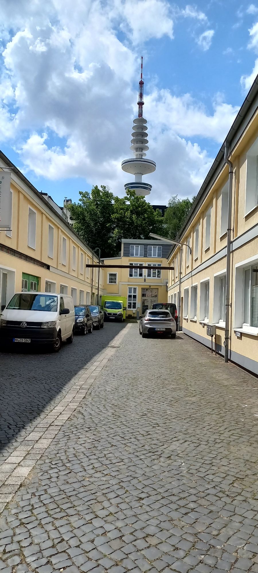 A cobblestone courtyard is flanked by yellow buildings, with vehicles parked along the sides. In the background, a tall tower is visible against a cloudy sky, providing a glimpse of the surroundings.