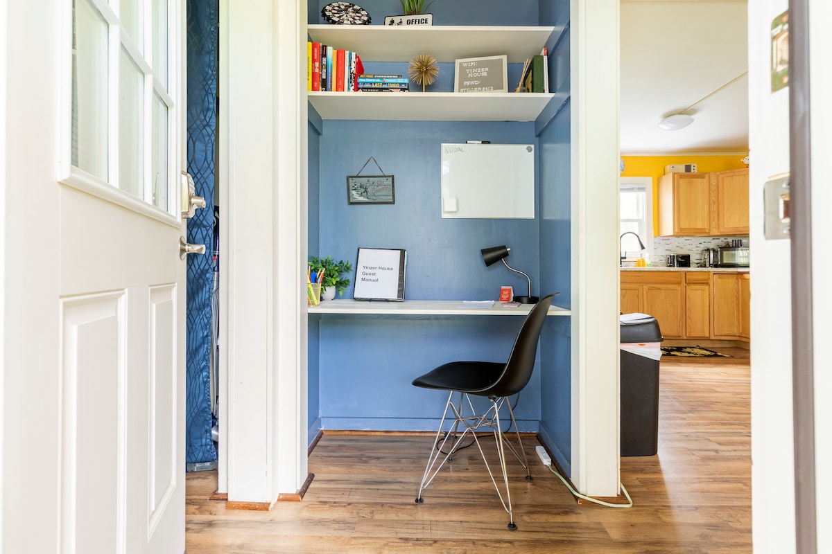 A small workspace is situated near the entrance, featuring a desk against a blue wall. A sleek chair is placed in front of the desk, which holds a whiteboard and small plants. Books are neatly arranged on a shelf above the desk.
