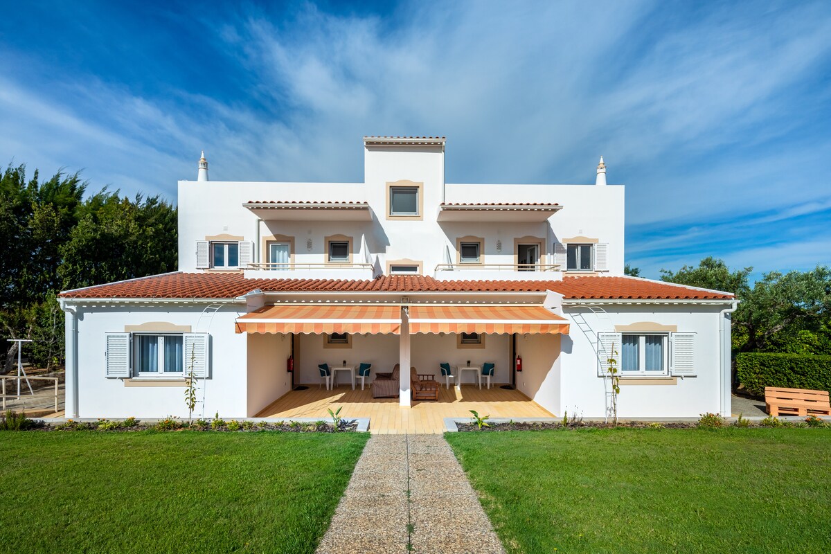 A white multi-unit building is presented, featuring a terracotta-tiled roof and multiple windows. A covered terrace with outdoor seating is visible at the front, complemented by a well-manicured green lawn and a gravel pathway leading to the entrance.