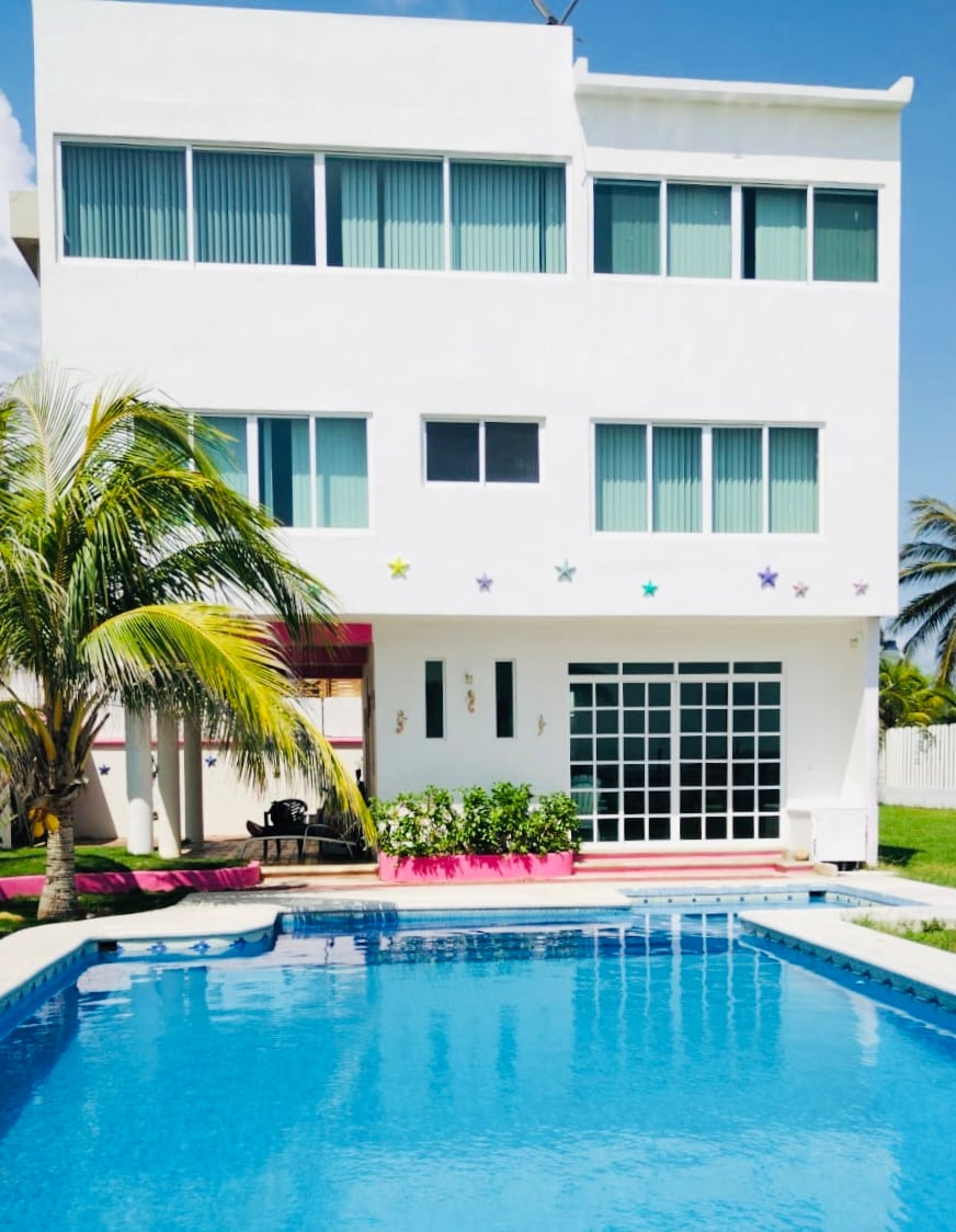 A three-story white building is set against a clear blue sky, featuring large windows that allow natural light. A clear pool is positioned in front, with a small border and lounge area visible. Lush greenery surrounds the property, enhancing the tropical atmosphere.