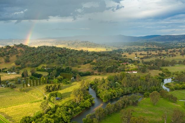 Aerial view showcases rolling hills and lush greenery surrounding the Tumut River. A subtle rainbow arcs across the sky, blending with the soft clouds above. The landscape features a mix of farmland and natural vegetation, highlighting the serene beauty of the Tumut region.