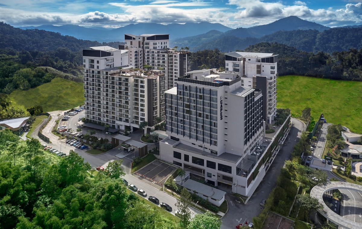 An aerial view showcases the modern architecture of the expansive apartment complex, surrounded by lush greenery and rolling hills. The structure features multiple buildings with balconies, offering a glimpse of the serene landscape in the background, under a partly cloudy sky.