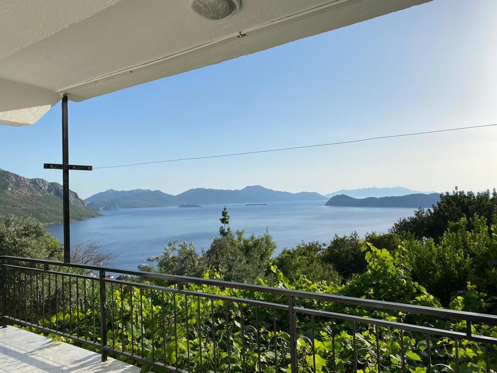 A balcony view features a panoramic landscape of calm waters and distant hills. Lush greenery frames the scene, enhancing the natural beauty of the coastal setting. The clear sky and gentle sunlight create a serene atmosphere, ideal for relaxation.