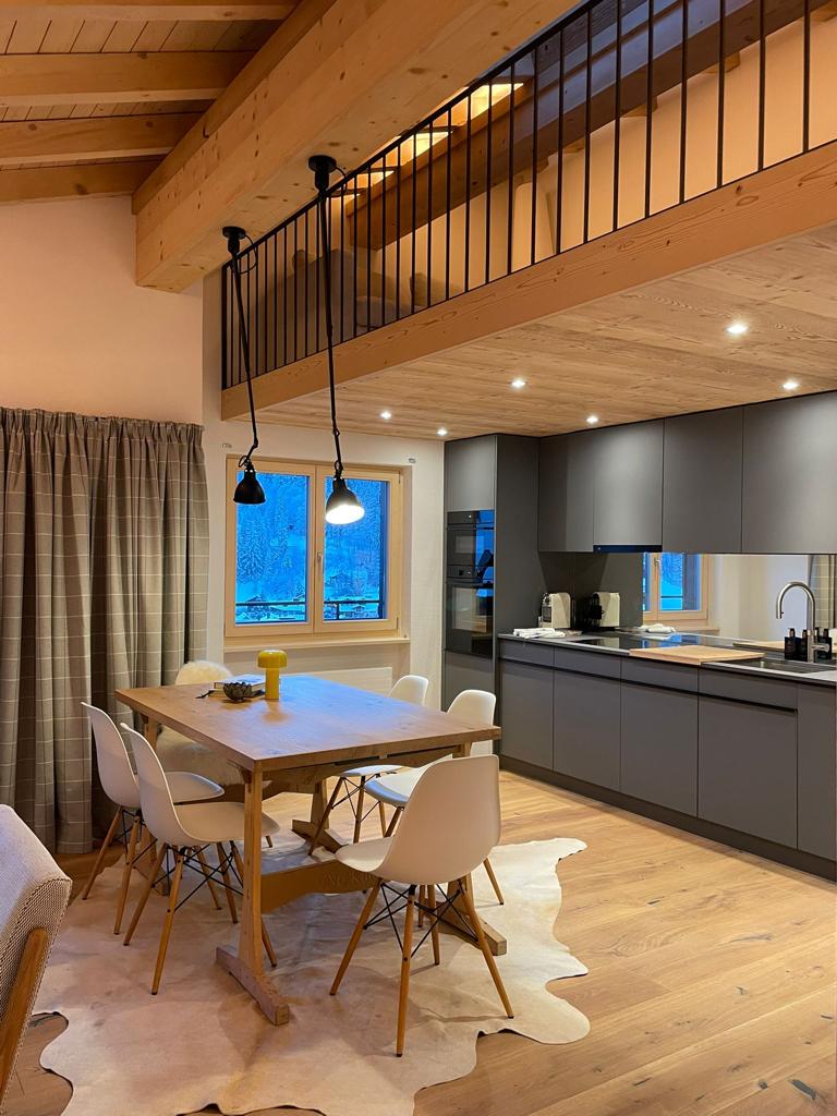 An open dining area features a light wood table surrounded by six white chairs. A modern kitchen is visible in the background with dark cabinetry and stainless steel appliances. Natural light enters through a large window, complemented by warm lighting from pendant fixtures.