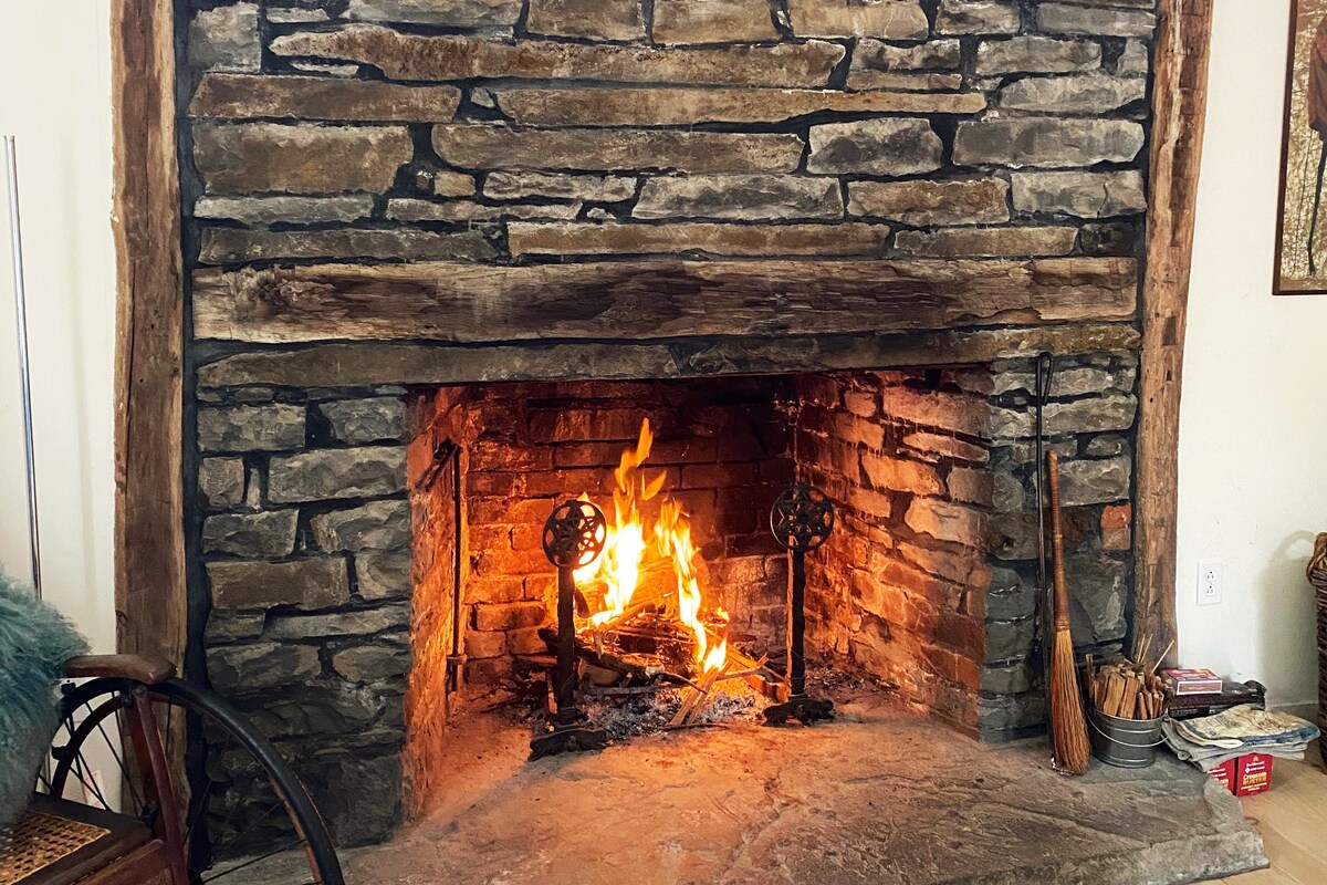 A large stone fireplace dominates the living area, featuring a warm fire crackling within. The rustic stonework encompasses the fireplace, adding texture to the space. A smooth stone hearth extends beneath, complemented by various tools nearby, creating a cozy focal point.