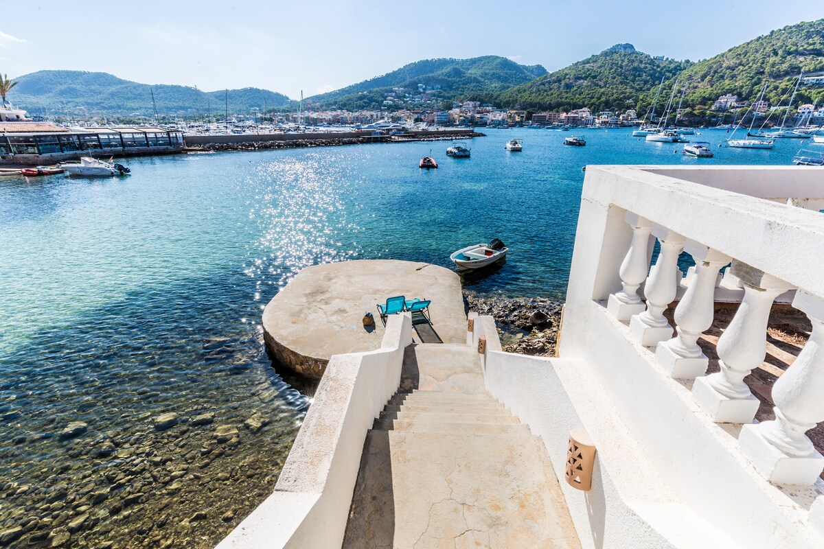 A staircase leads down to a small, circular platform by the water's edge, with views of the clear blue sea. The platform is surrounded by pebbles and small boats are visible in the distance. Sunlight sparkles on the water's surface, enhancing the coastal scenery.