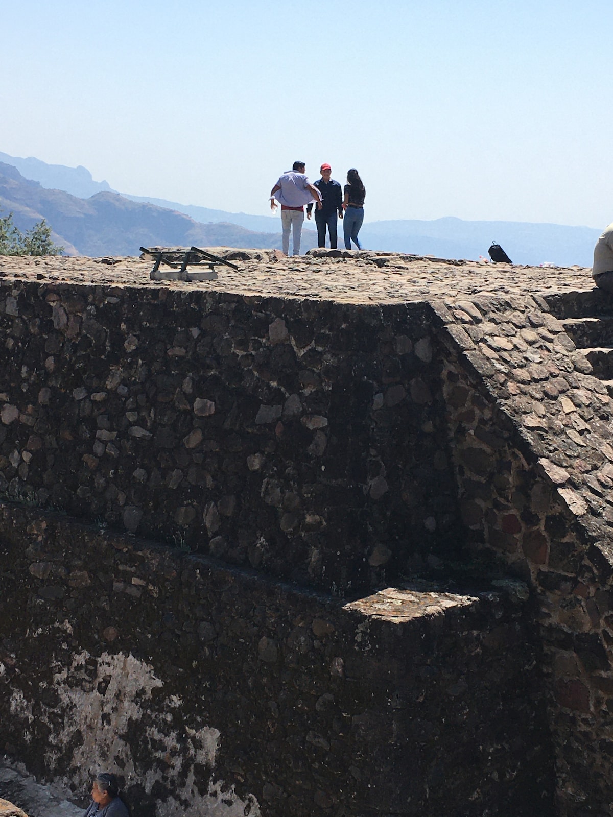 A group of individuals stands on top of a stone structure with a view of distant mountains and a clear blue sky. The stone surface exhibits a mix of rough textures, emphasizing the historic nature of the site.
