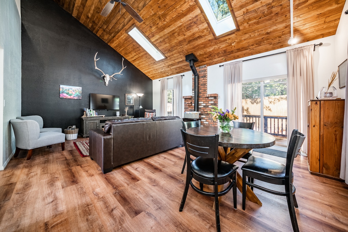 A spacious living area features a vaulted wooden ceiling with skylights allowing natural light to enter. A comfortable couch faces the TV and a round dining table is situated nearby. Large windows provide a view of the outdoor patio, with neutral-colored walls enhancing the inviting atmosphere.