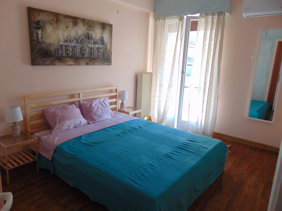 A light-filled bedroom features a queen bed dressed in a turquoise coverlet, complemented by soft pink pillows. A mirror reflects the space, while a sliding glass door opens to a small balcony. Wooden flooring adds warmth to the inviting room.