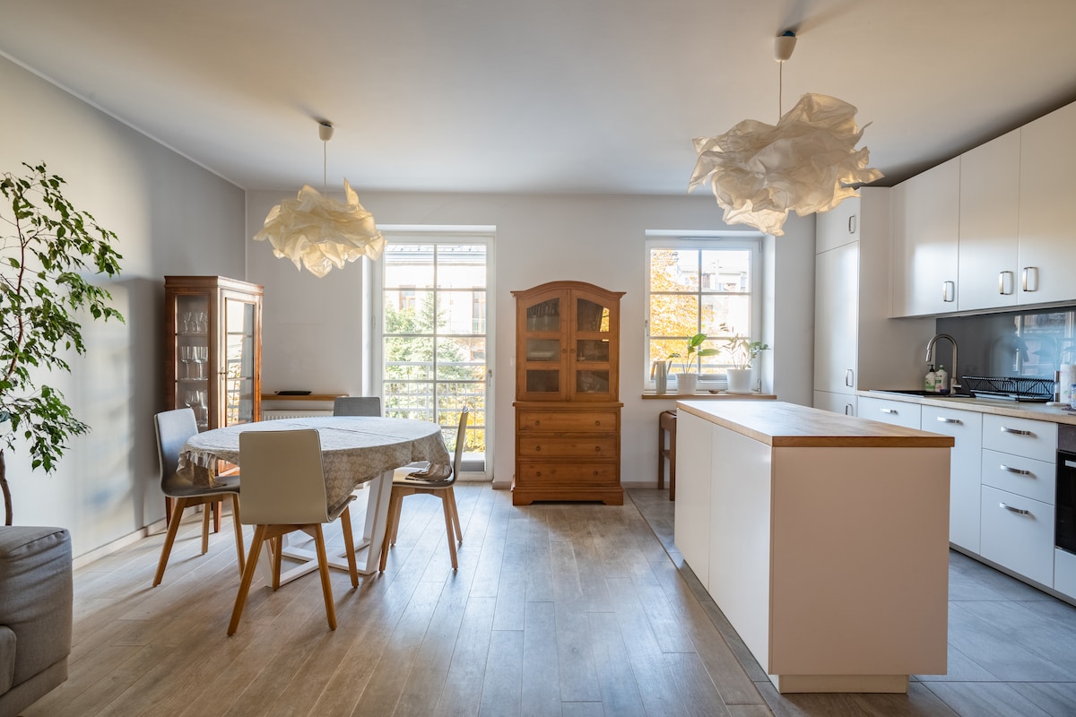 A bright dining area is complemented by large windows allowing natural light to fill the space. A circular table with four chairs is positioned near a wooden china cabinet. The modern kitchen features white cabinetry, an island with a wooden countertop, and minimalistic pendant lighting.