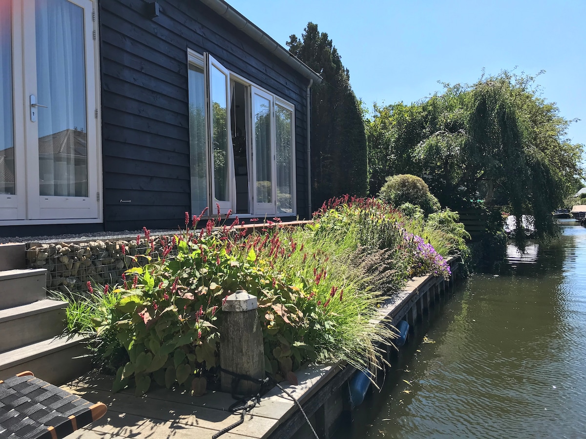 The exterior of the house is visible, showcasing a wooden structure with large glass doors. A garden filled with vibrant flowers and greenery spreads along the pathway, leading to a deck by the water, where plants add natural beauty to the landscape.