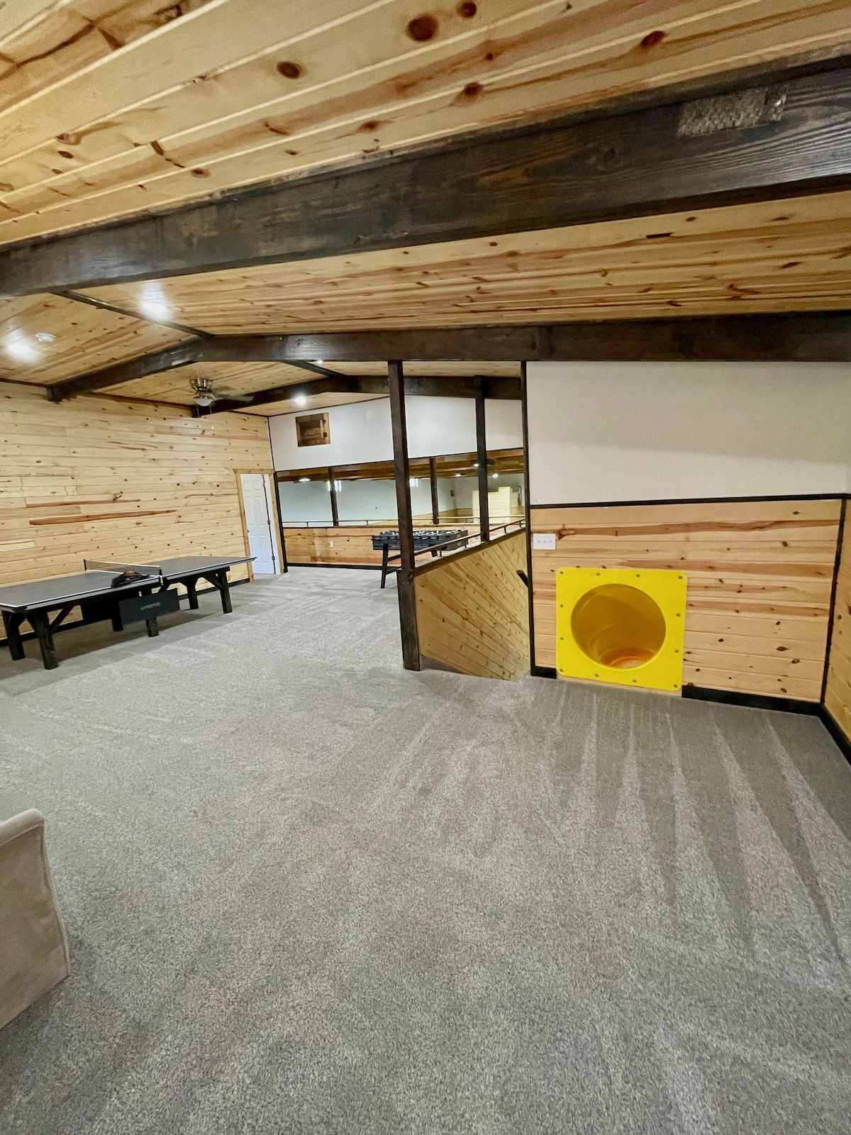 A spacious game room is showcased with wooden paneling and a carpeted floor. A yellow play tunnel adds a playful touch. Various game tables are positioned for fun activities, and natural light enters through windows, enhancing the overall inviting ambiance.