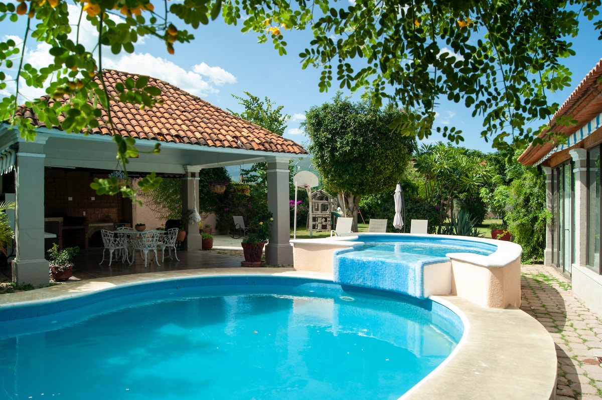 A refreshing outdoor pool area features two interconnected pools, surrounded by lush greenery and colorful plants. A shaded gazebo with white wicker furniture provides a relaxing space, while a built-in jacuzzi is positioned next to the main pool. Sunlight filters through the trees, creating a serene ambiance.