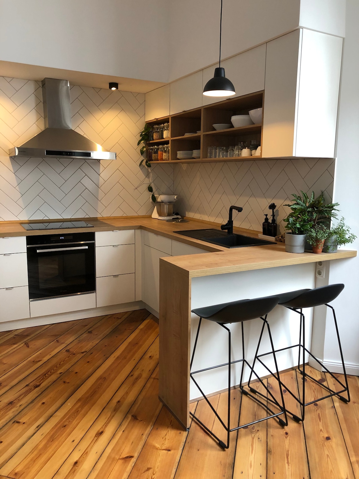 A modern kitchen features a compact design with a combination of sleek cabinetry and natural wood accents. The space includes a built-in oven, a stylish black sink, and bar stools at a small countertop. Soft lighting highlights the herringbone-patterned backsplash.