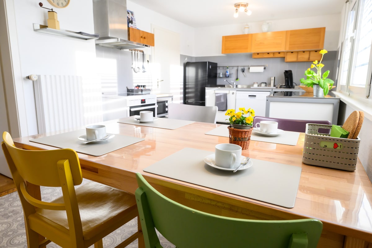 A spacious kitchen area is shown with a wooden table set for four, each place featuring a cup and saucer. Colorful chairs and a small potted plant with yellow flowers add warmth. Kitchen appliances and cabinetry are visible in the background, creating a functional cooking space.