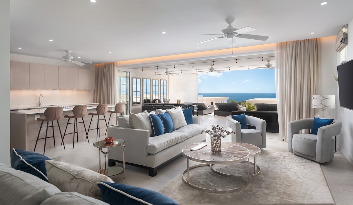An open-plan living area is featured, showcasing a modern design with comfortable seating arrangements. Large windows frame expansive sea views, and a sleek kitchen bar with stylish stools is visible in the background. Soft neutral tones are complemented by hints of blue accents.