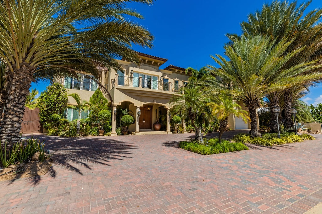 The villa's exterior is showcased with a sprawling brick driveway lined with palm trees and tropical plants. A grand entrance features arches and decorative elements, creating an inviting approach to the residence under a clear blue sky.