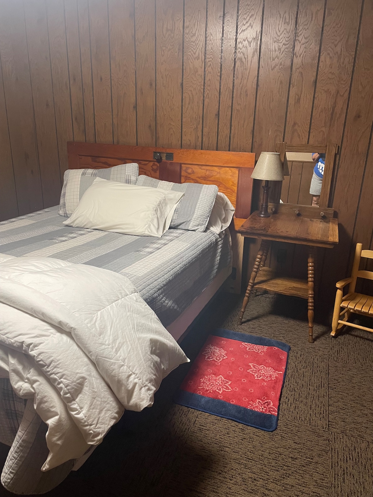 A cozy bedroom features a wooden bed with a patterned quilt and three accent pillows. A small wooden table with a lamp and a mirror complements the space, while a plush blue rug adds warmth beneath the bed. A child's wooden chair offers additional seating.