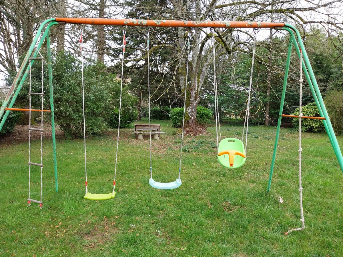 A children's swing set is positioned on a grassy area, featuring three swings in various colors: a green swing, a light blue swing, and a single, yellow toddler seat. A wooden bench is visible in the background, surrounded by trees and bushes.