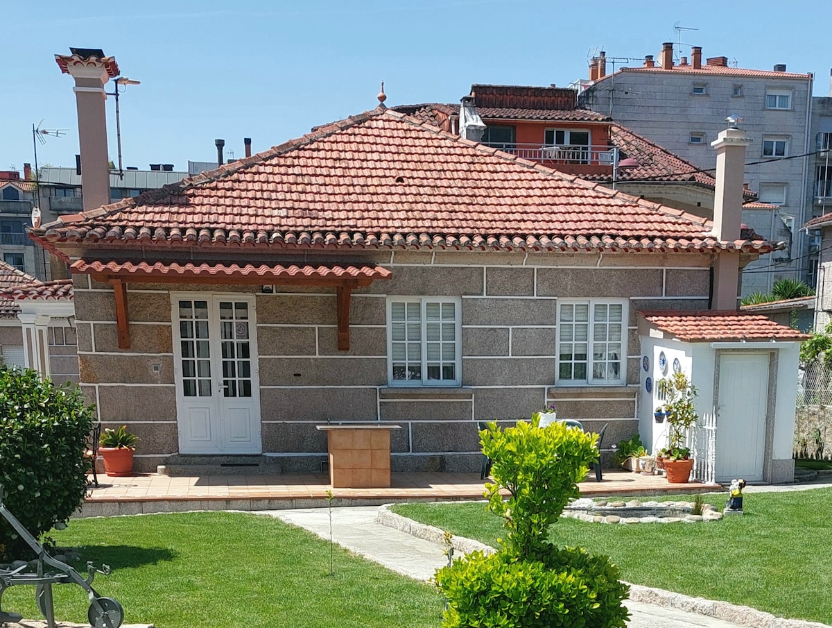 The charming stone house displays a tiled roof and prominent front porch. Twin white doors and window frames enhance the façade, while potted plants add a touch of greenery. A well-kept garden with edged pathways leads to the entrance, surrounded by residential buildings in the background.