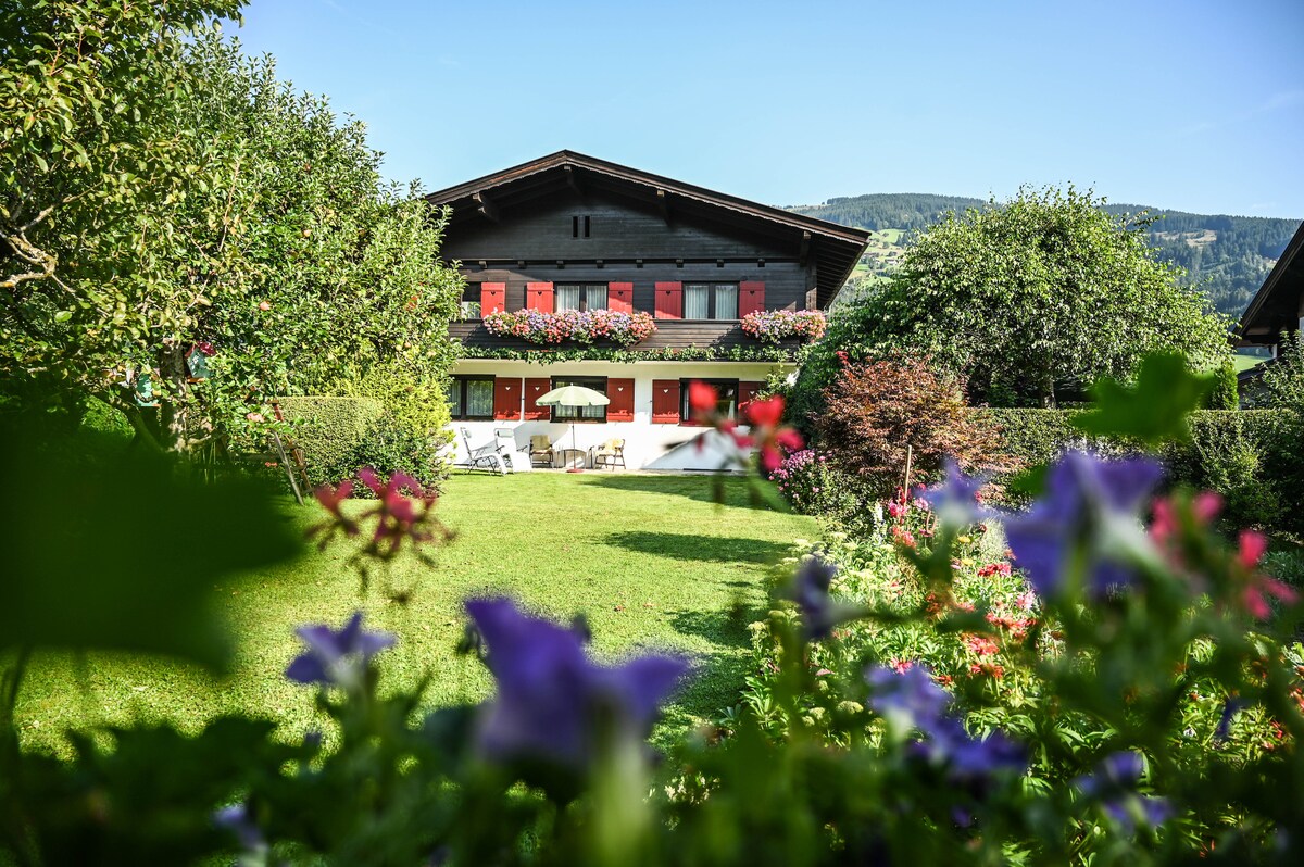 A traditional chalet-style building is framed by lush greenery and vibrant flowers in the foreground. The exterior features wooden balconies adorned with flower boxes, while a patio area with seating is visible below. The backdrop includes rolling hills under a clear blue sky.