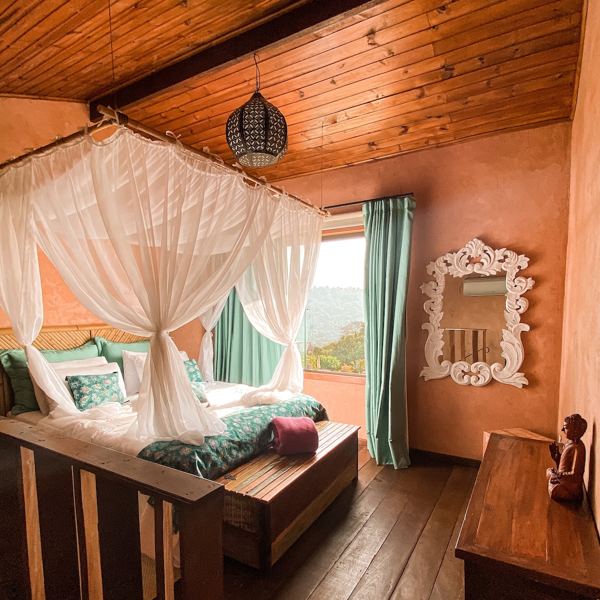 A cozy room features a spacious bed draped with sheer white fabric, offering a calming ambiance. Green curtains frame a window with a mountain view. A decorative mirror and a wooden dresser complement the warm, earthy tones of the walls and wooden flooring.