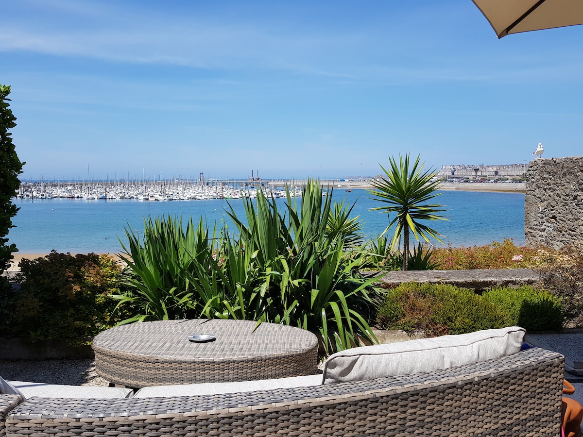 A view of the marina can be seen from a seating area, featuring a round wicker table surrounded by comfortable cushions. Lush plants frame the scene, with the sea in the background and clear blue skies above.