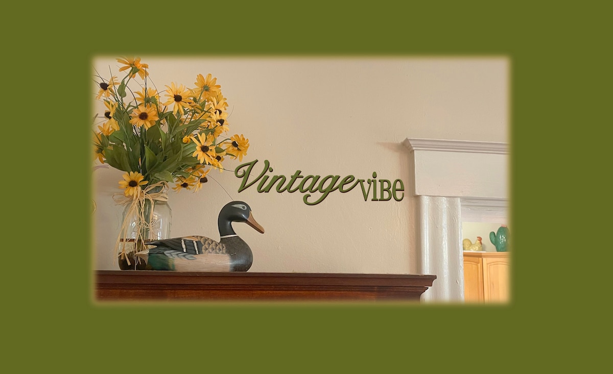 A decor piece featuring a ceramic duck is displayed atop a wooden surface. A glass vase filled with bright yellow flowers adds a touch of warmth. The word 'Vintage' is artistically positioned on the wall, complemented by a soothing green backdrop.